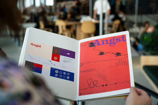 Hender som holder en bok åpen. Sidene viser typografi og andre grafiske elementer. Foto