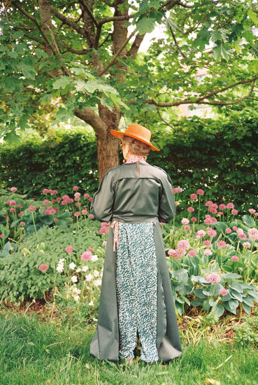Eldre dame står ute i en hage, kledd i mørk og lang kjole og oransje hatt. Foto