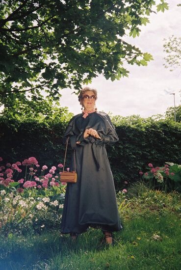 Eldre dame står smilende ute i en hage, med mørk kjole, solbriller og lys brun veske. Foto