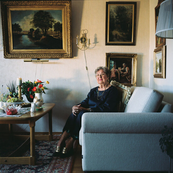 Eldre dame sitter smilende vendt mot kamera i en sofa. Foto