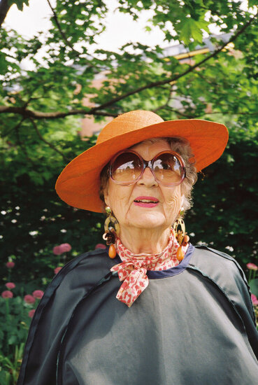 Nærbilde av eldre dame med solbriller, oransje hatt og mørk kjole. Foto