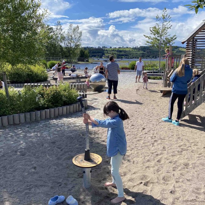 Lekeplass med dekke av sand med barn som leker og voksne som ser på. 