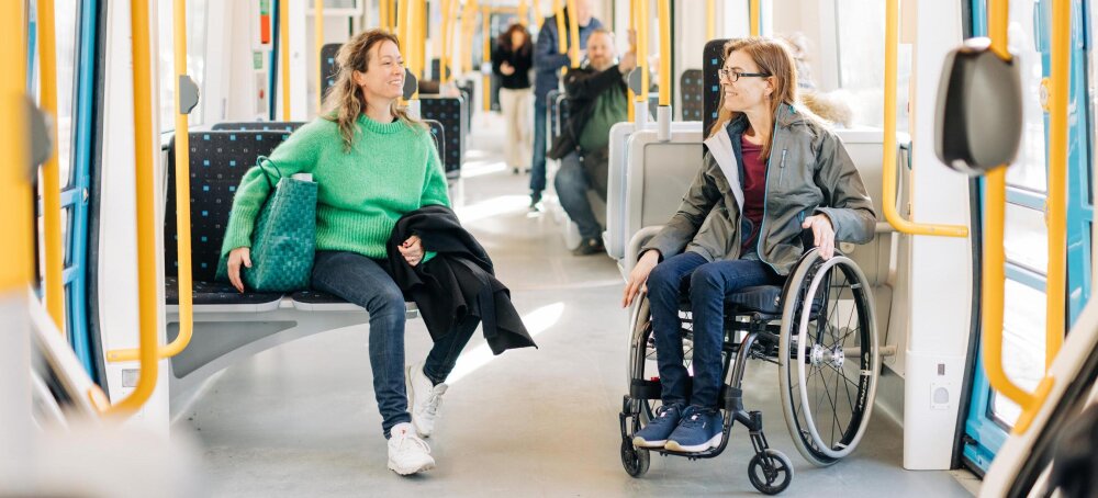 To kvinner sitter på hver sin side av midtgangen på trikken. Den ene sitter på et sete, den andre sitter i rullestol. De ser på hverandre og smiler. 