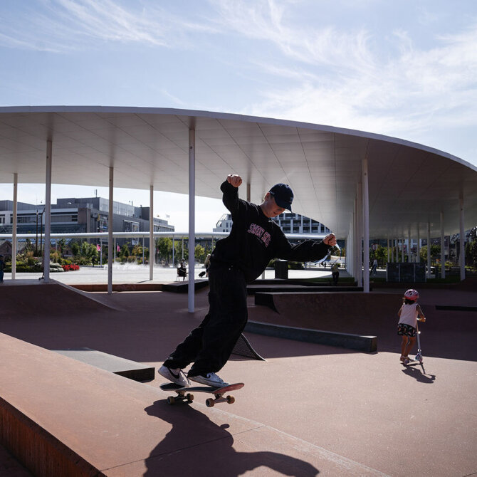 En ung mann skater på en skatebane. I bakgrunnen, et barn på sparkesykkel.