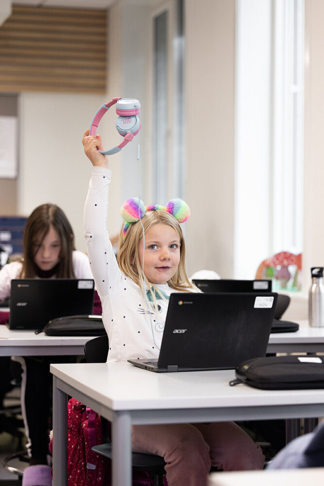 En jente rekker opp hånda og sitter i et klasserom med en bærbar pc foran seg. Foto