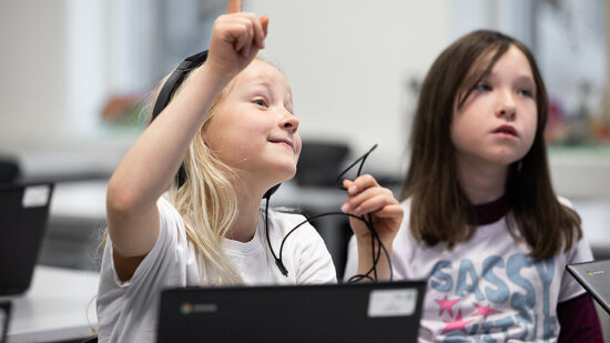 To jenter i et klasserom som rekker opp hånda. Foto