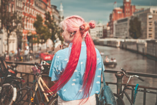 Jente med rosa hår som står på en kanalbro i Amsterdam. Sykler er lent opp mot rekkverket på broen.