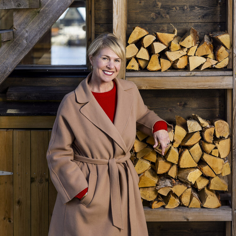 En smilende kvinne lent opp mot en vedstabel. 
