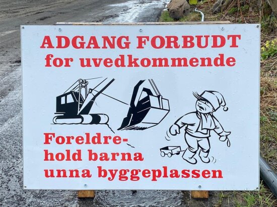 Skilt med teksten adgang forbudt i rødt og med store bokstaver øverst. Deretter for uvedkommende . Under en svart hvitt-illustrasjon av et barn stående foran en gravemaskin. Under illustrasjonen er det mer tekst i rødt der det står Foreldre – hold barna unna byggeplassen. Bak skiltet skimtes en sølete anleggsvei.