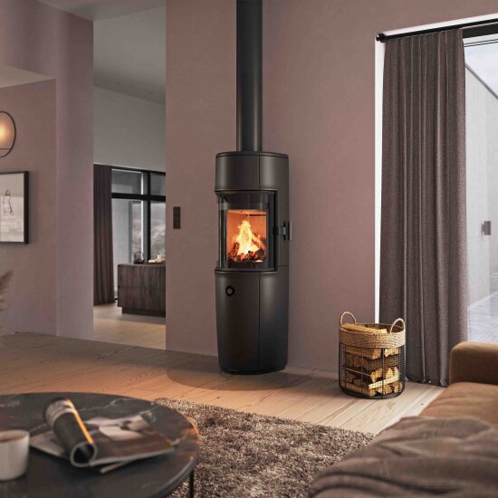 En stue i lune farger , langlugget teppe på gulvet, og en kurv med ved. Midt i rommet brenner det et stort peisbål i en frittstående, sort peis med glassdør og sort rør opp til taket.