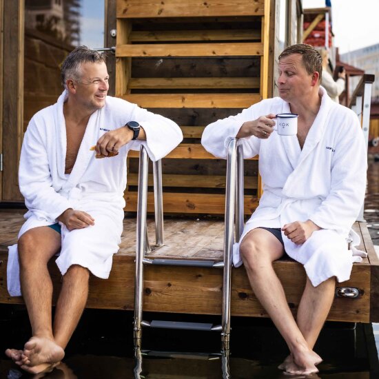 To menn i hvit badekåpe sitter foran en badstue med føttende dinglende i vannkanten.