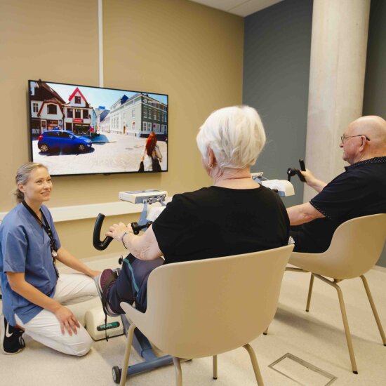 To eldre mennesker sitter og ser på en skjerm mens de holder på et slags styre. En helsearbeider sitter på kne ved siden av dem og følger med.