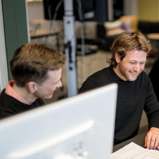 To unge menn sitter ved siden av hverandre og ser smilende på en skjerm.