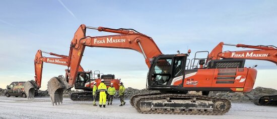 Fire menn i gult arbeidstøy atår under grabben på en stor rød gravemaskin.
