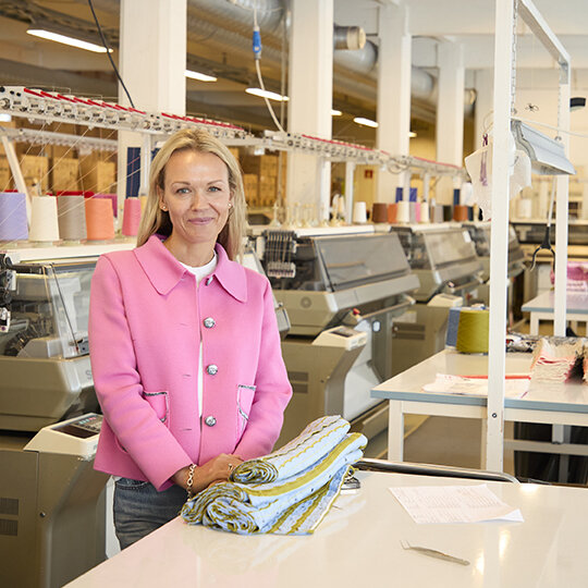 En smilende dame med rosa jakke foran strikkemaskiner og garn.