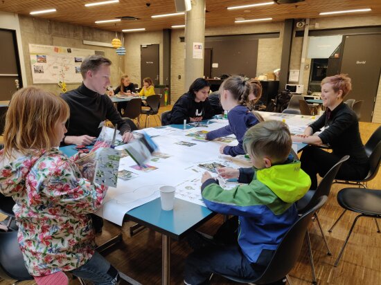 Barn på workshop sammen med voksne gruppeledere. De sitter rundt et bord og holder på med ark og bilder.