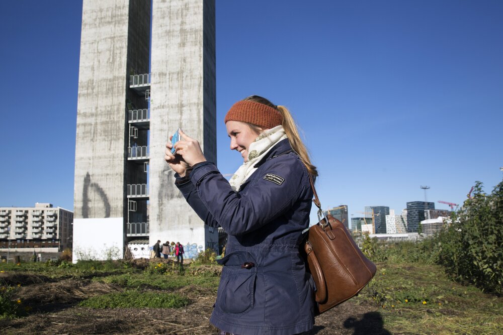 Kvinne med blå parkas og strikket ørevarmer tar bilder med mobilkamera. Stor, grå ventilasjonspipe i bakgrunnen.