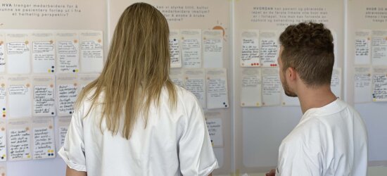 En man og en kvinne i hvite sykehusklær, med ryggen til fotografen, studerer en tavle med håndskrevne lapper.