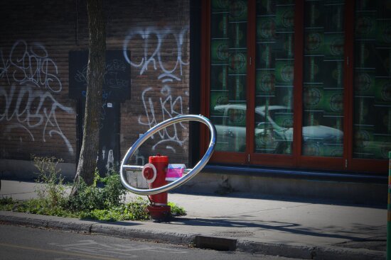 En brannhydrant med en stor ring påmontert. Ut av ringen kan det komme drikkevann. I bakgrunnen, murvegger med tagging og vindusruter som reflekterer en bil.