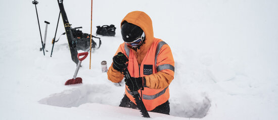 Bildet viser en person som står i dyp snø med en snøsag i hendene. i Bakgrunnen ski og staver.
